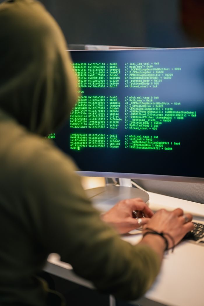 hero-img-01 Person wearing a hoodie typing code on a computer screen with green text, depicting hacking scene.